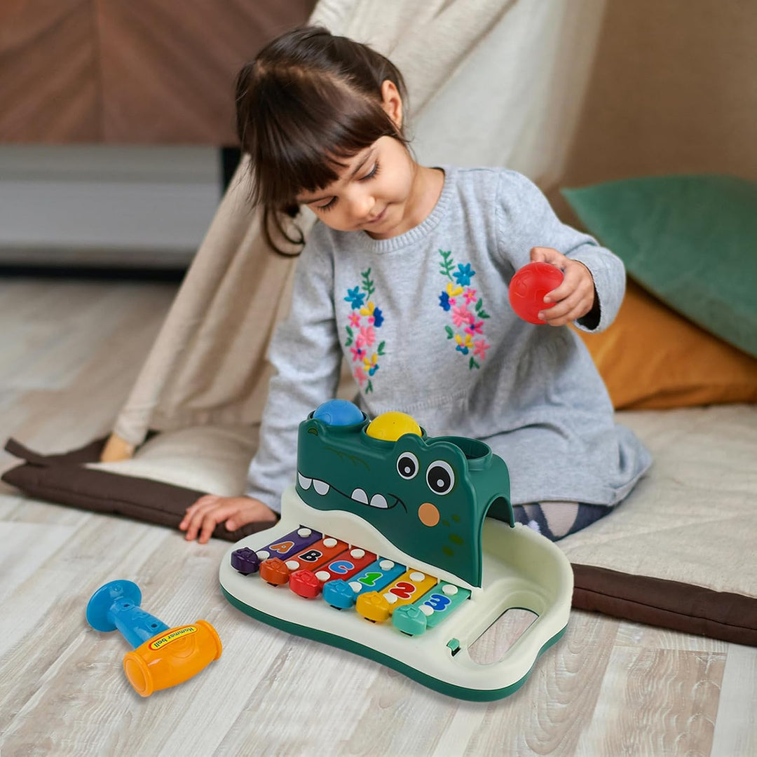 Child holding a red ball beside the alligator xylophone toy on the floor