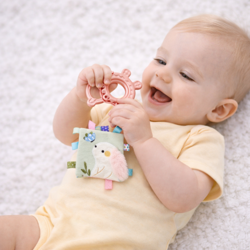 Baby holding a pink teething ring attached to a textured cloth book