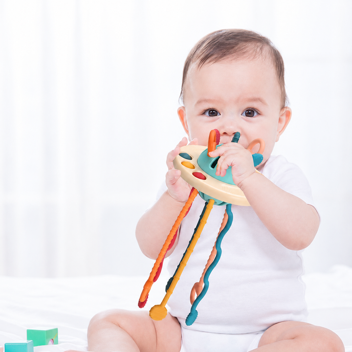 Baby holding a UFO-style sensory toy near their mouth, with textured cords hanging below