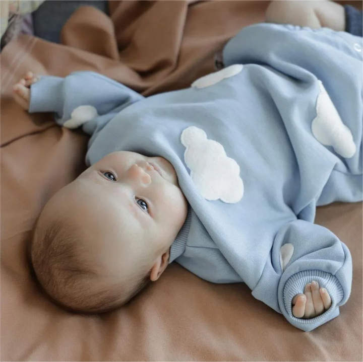 Baby lying on a blanket in a blue long-sleeve romper with white cloud appliques.