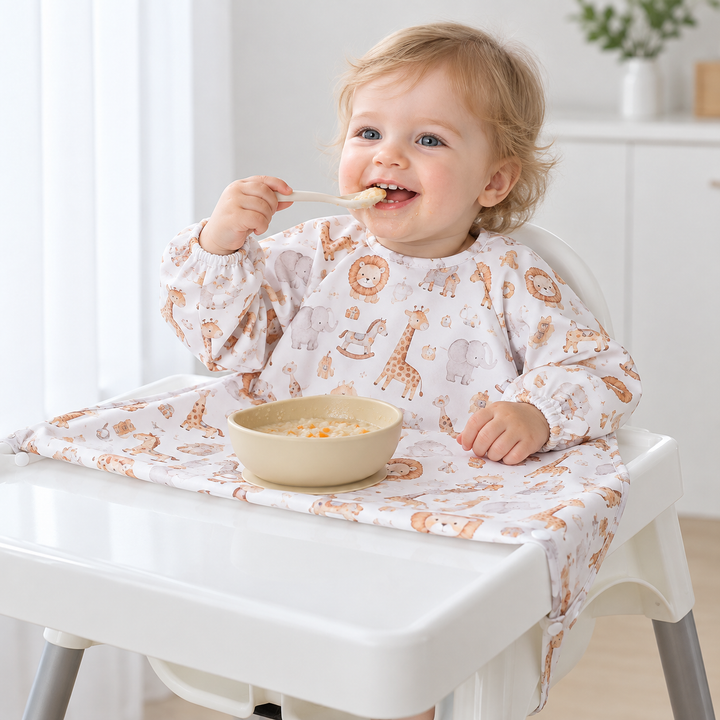 Baby using a spoon while wearing a long-sleeve animal-print bib over a high-chair tray