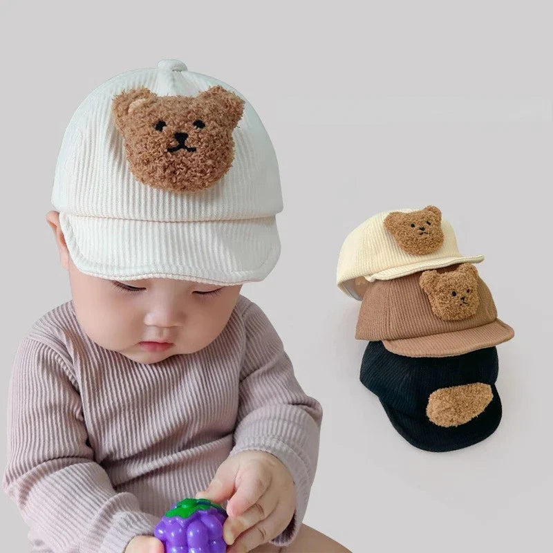 Baby wearing a white corduroy bear cap beside a stack of white, brown, and black caps