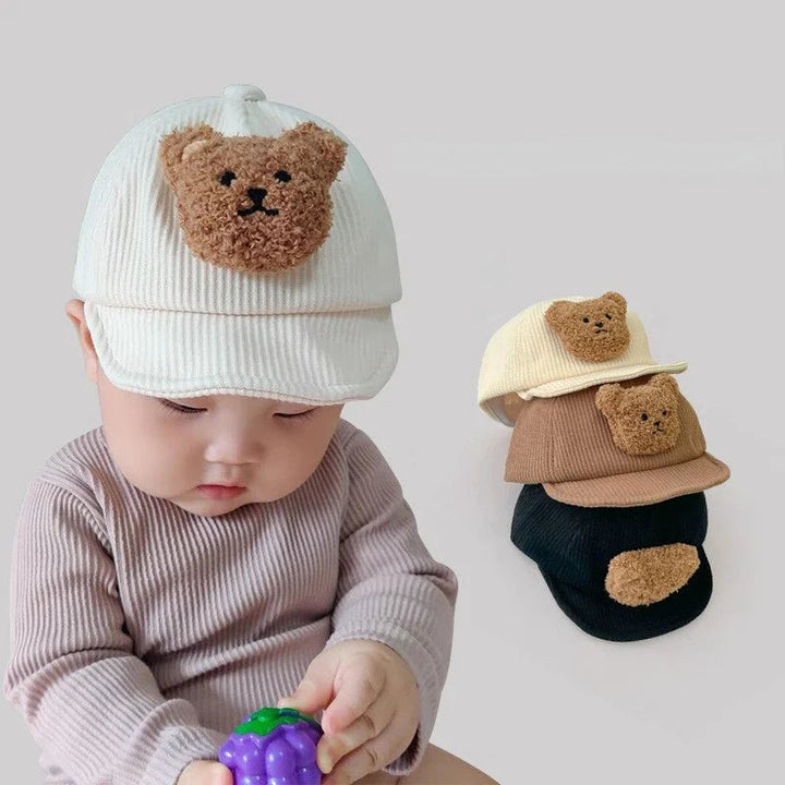 Baby wearing a white corduroy bear cap beside a stack of white, brown, and black caps