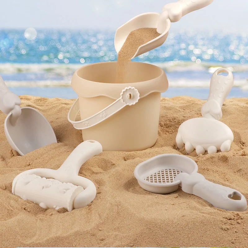 Sand pouring from a scoop into a beach bucket surrounded by a shovel, rake, sieve, and mold