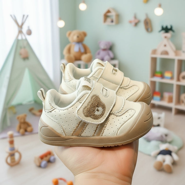 Hand holding a pair of cream bear-detail sneakers in a nursery setting