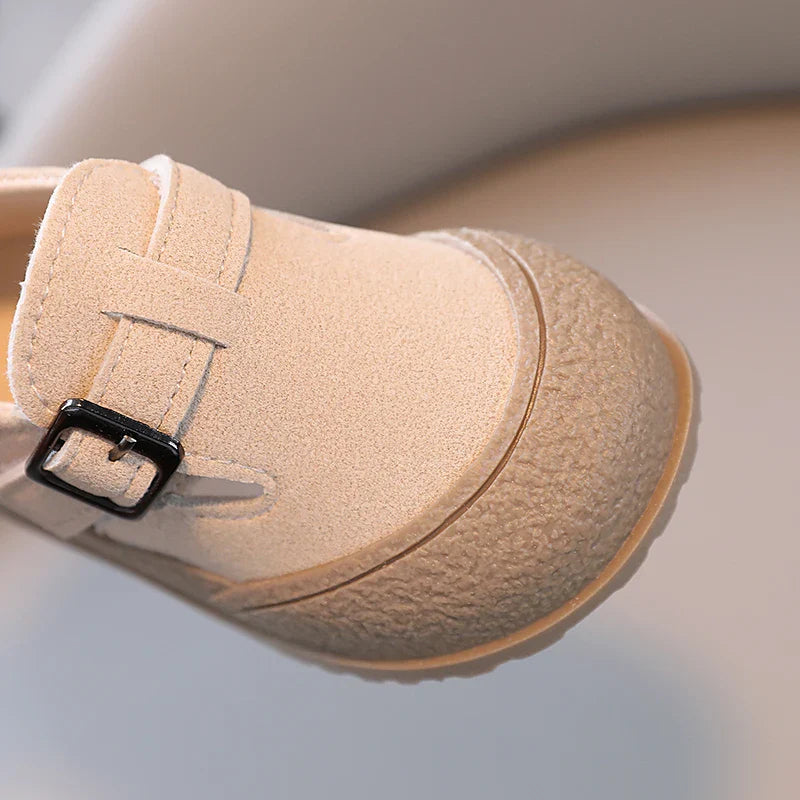 Close-up of a beige baby shoe with a black buckle and textured toe edge