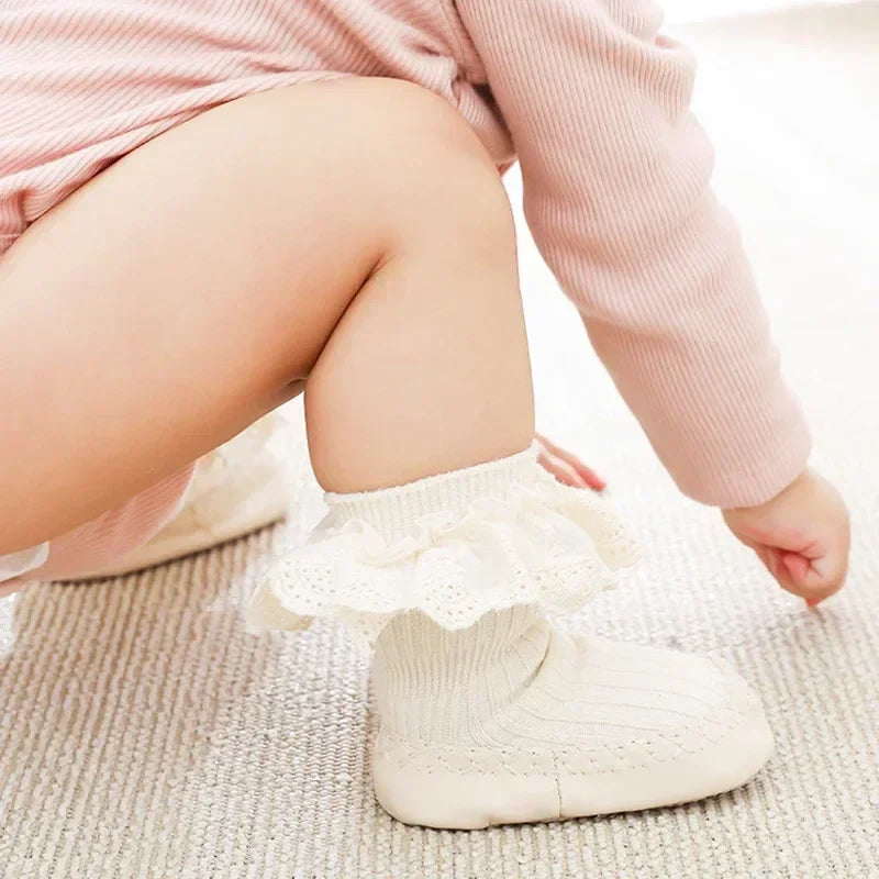 Baby wearing beige sock shoes with cream lace cuffs while crouching on a rug