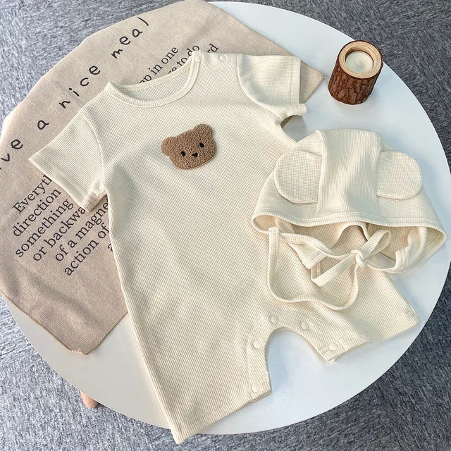 Beige cotton baby romper with bear chest detail and matching hat on a round table
