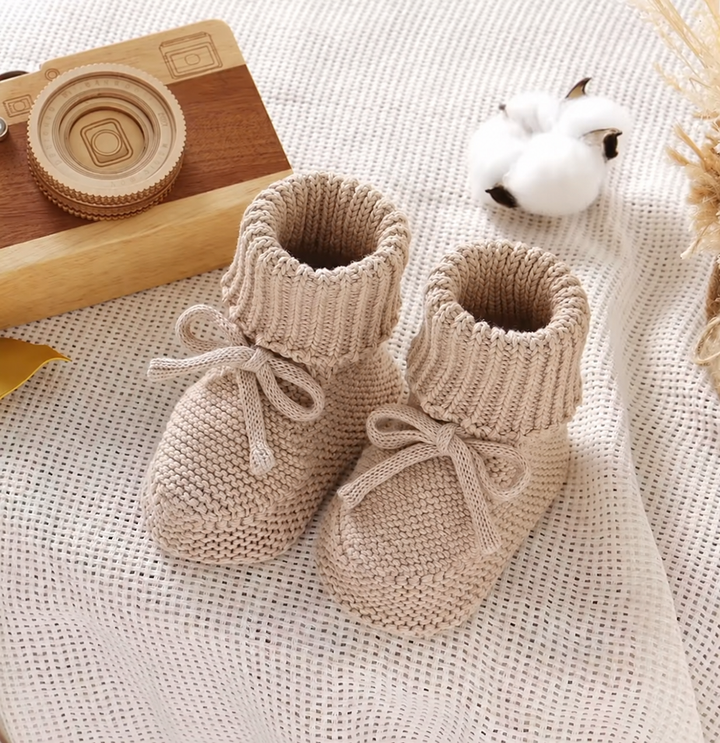 Beige cotton-wool baby booties on a knit blanket beside a wooden toy camera