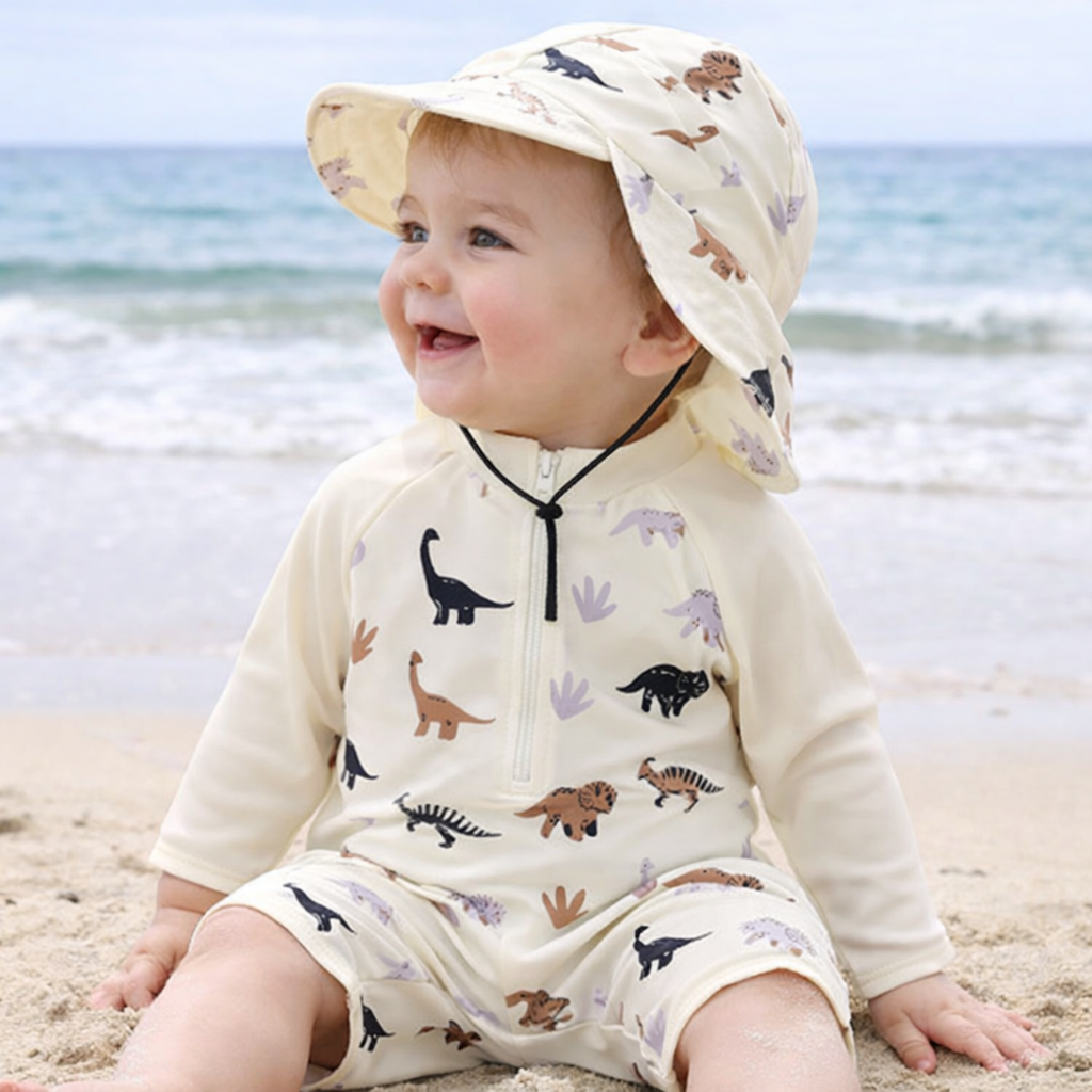 Baby wearing a beige dinosaur-print swimsuit and matching wide-brim hat at the beach.