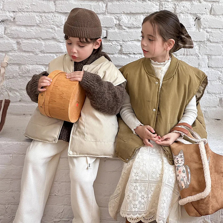Children seated in beige and olive green vests against a white brick wall.