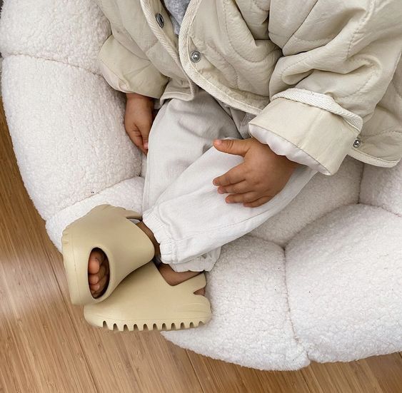 Child in a neutral outfit wearing beige sandals while sitting on an upholstered chair.