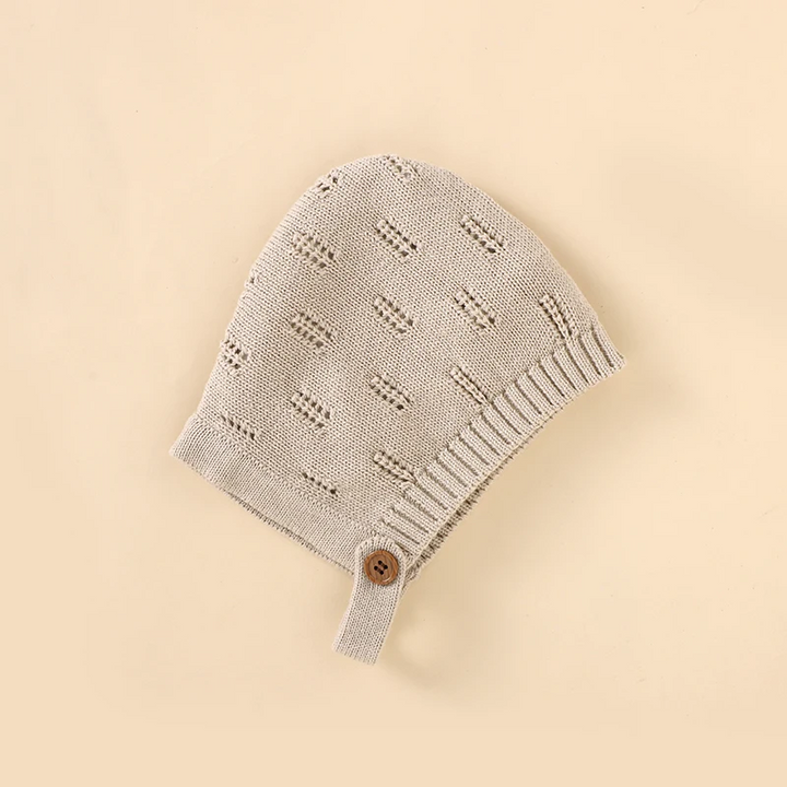 Beige knitted baby hat showing the patterned knit, ribbed edge, and button tab.