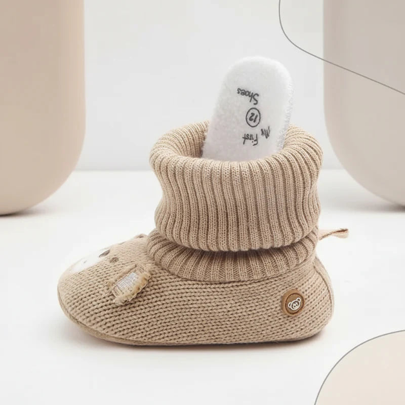 Side view of a beige knit baby bootie with a folded cuff and white insert in the opening.