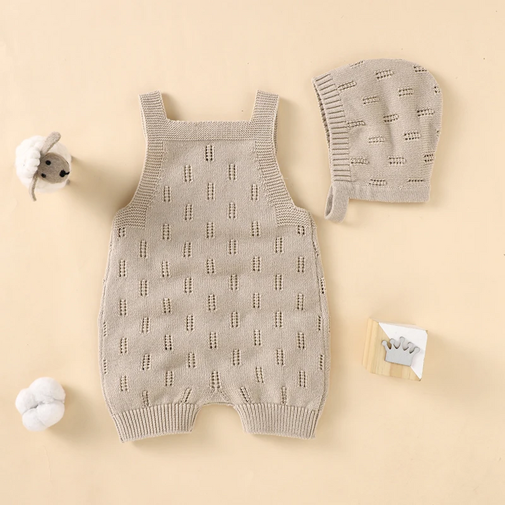 Beige knitted baby overalls and matching hat laid flat beside small nursery toys.