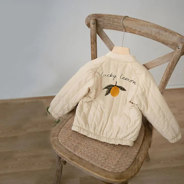 Beige quilted winter coat draped over a wooden chair, showing lemon embroidery on the back
