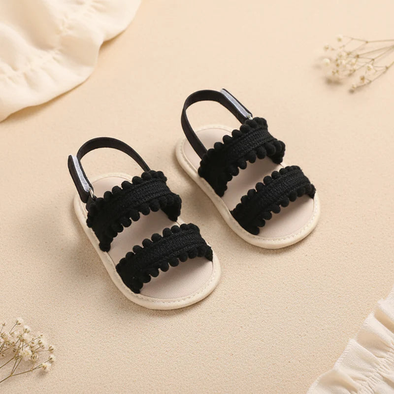 Black baby sandals with pom-pom-trimmed straps and contrasting light-colored soles.