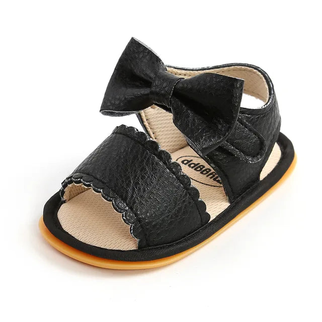 Black baby sandal with a bow, scalloped front strap, and open toe