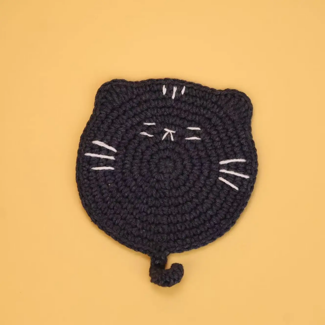 Black crocheted cat coaster with white stitched whiskers and a curled tail