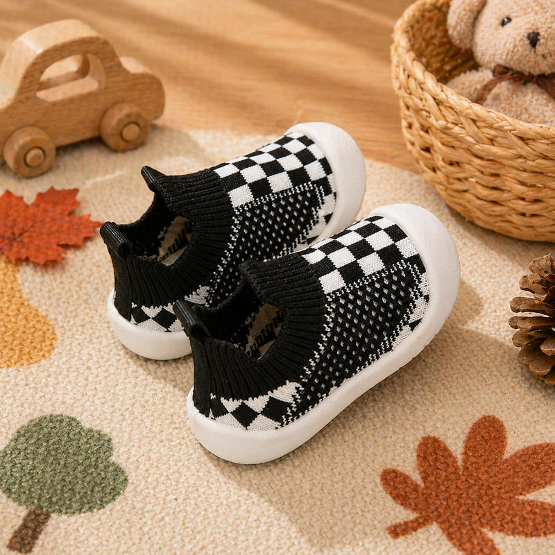 Black checkerboard baby sock shoes with ribbed cuffs and white soles on a patterned rug.