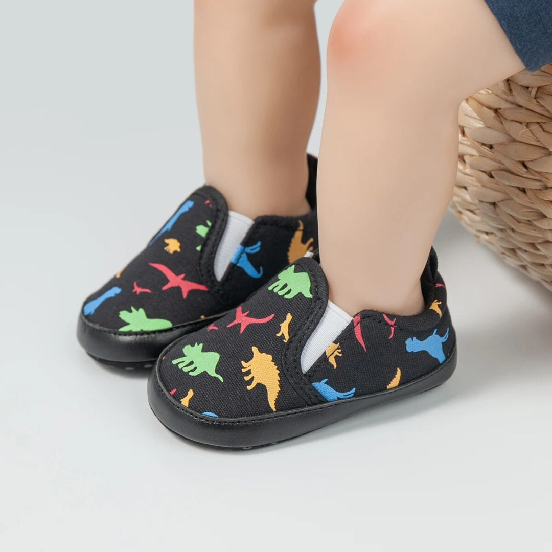 Black baby sneakers with colorful dinosaur prints and elastic side panels, shown on feet