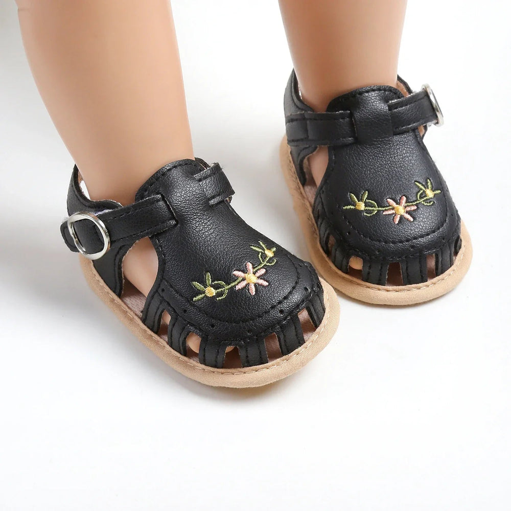 Black floral baby sandals with adjustable straps and cutout toes on mannequin feet