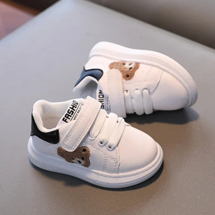 A pair of white bear sneakers with black heel accents and strap closures on a gray seat.