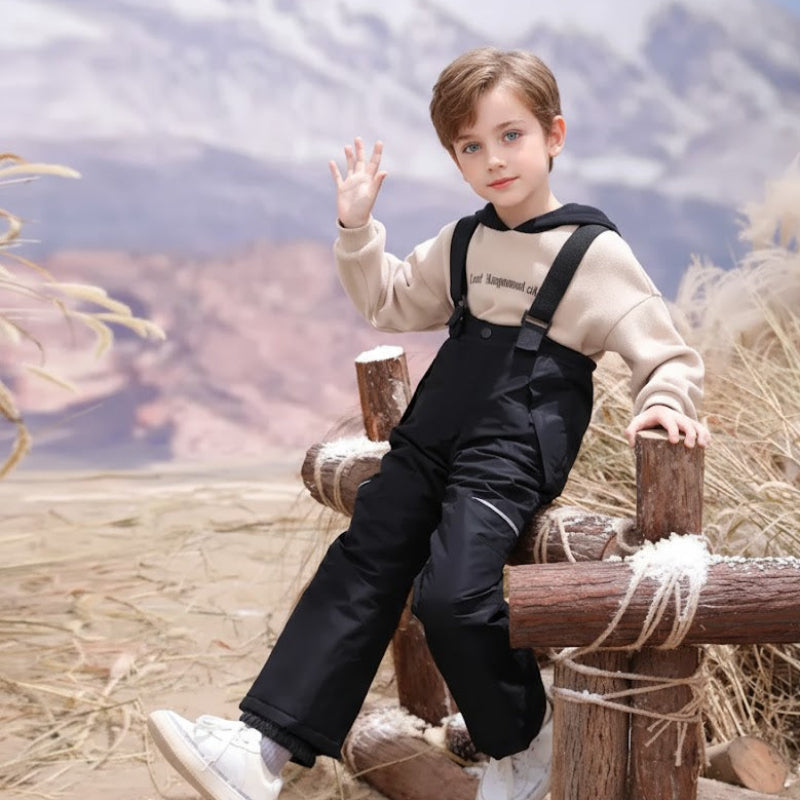 Child seated on a wooden fence wearing black ski pants with suspenders.