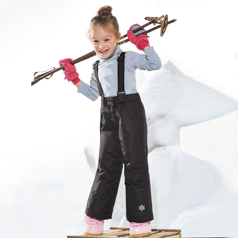 Child wearing black ski pants with suspenders and pink gloves in a snowy setting.