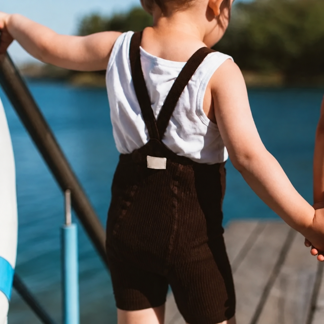 Back view of a child wearing black ribbed suspender shorts over a white tank top on a dock.