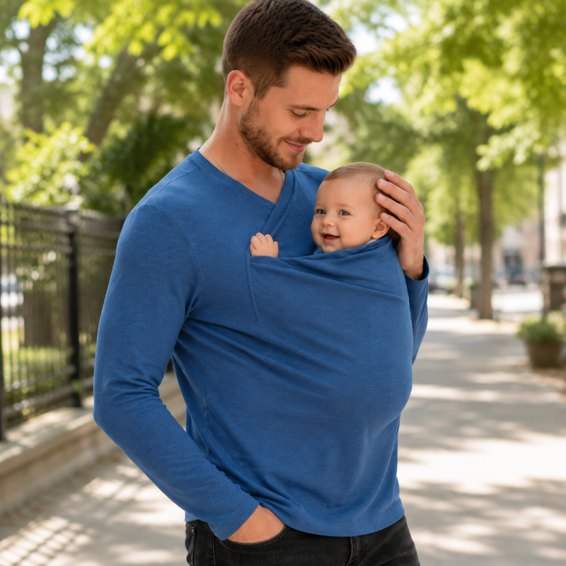 Man wearing a blue baby carrier shirt with a baby in the front pouch on a tree-lined path.