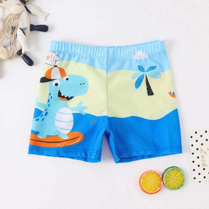 Blue swim trunks with a cartoon dinosaur on a surfboard and a beach scene.