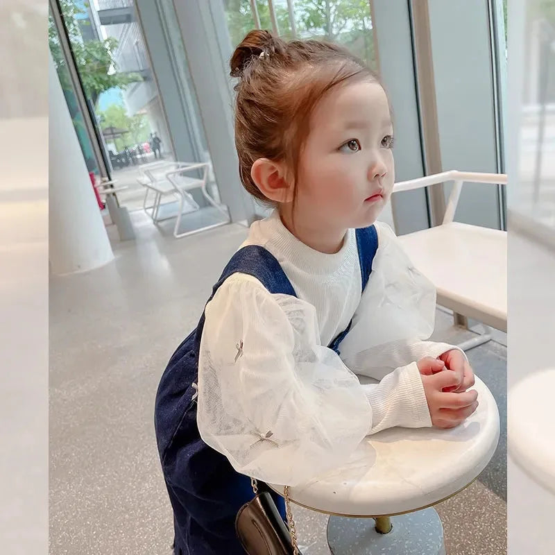 Child leaning on a table in blue overalls and a white long-sleeve blouse