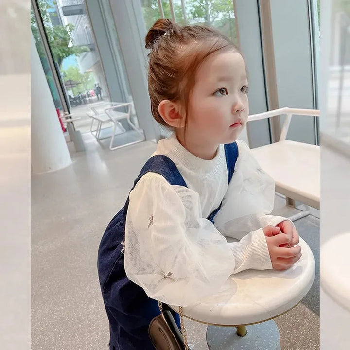 Child leaning on a table in blue overalls and a white long-sleeve blouse