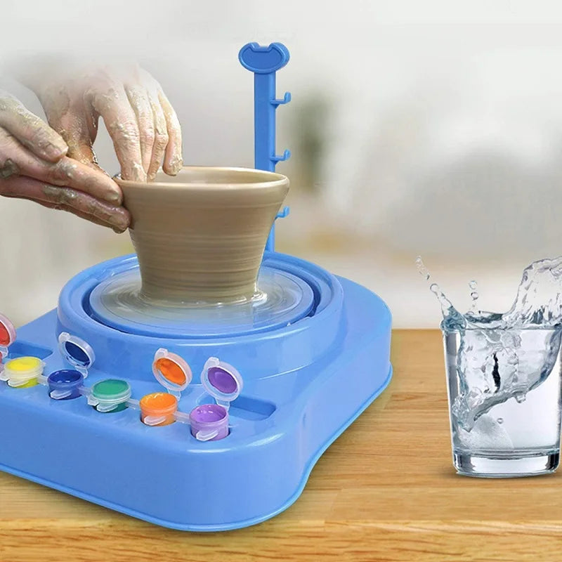 Hands shaping a clay bowl on a blue pottery wheel beside paint pots and a glass of water