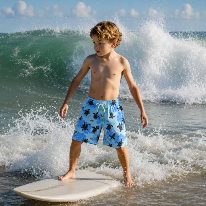 Boy wearing light blue turtle-print swim trunks while standing on a surfboard at the beach