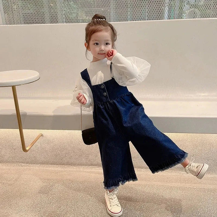 Child lifting one leg to show the roomy, wide-leg cut of blue overalls