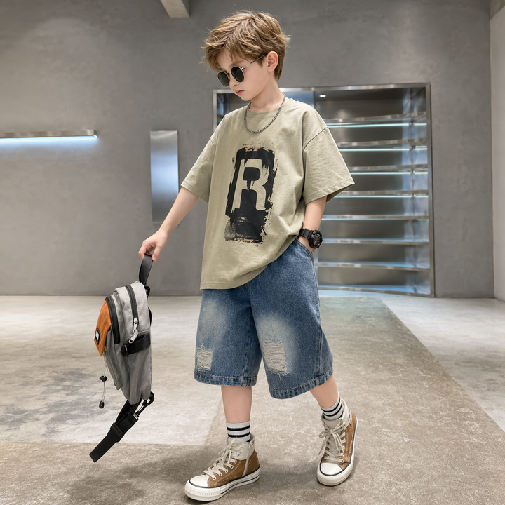 Boy wearing a beige R-graphic T-shirt and blue denim shorts.