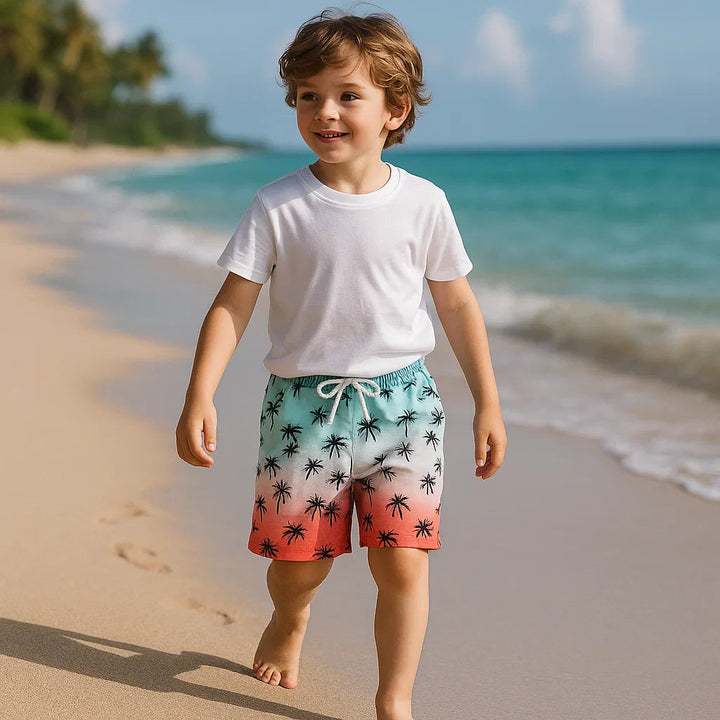 Boy wearing mint-to-coral palm-print swim trunks on a sandy beach.