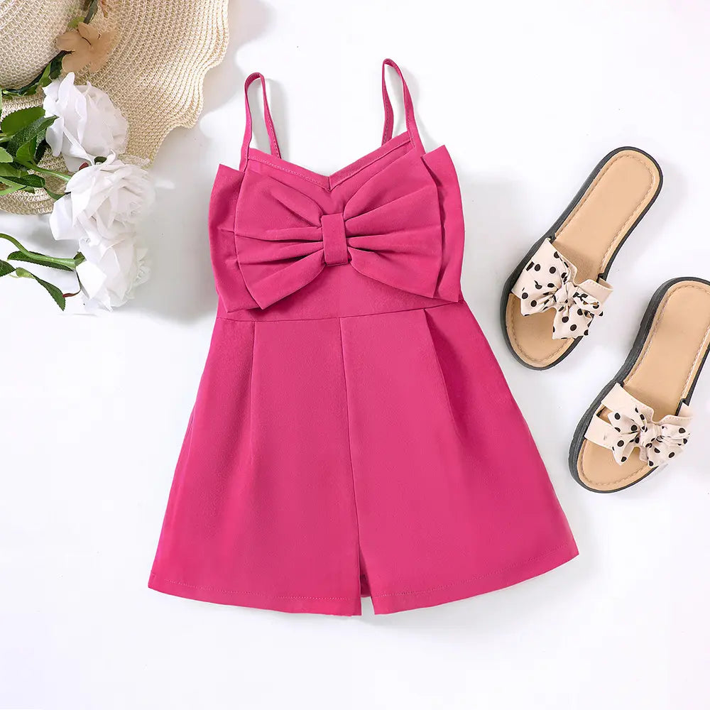 Bright pink bow-front romper laid flat beside sandals, white flowers and a straw hat