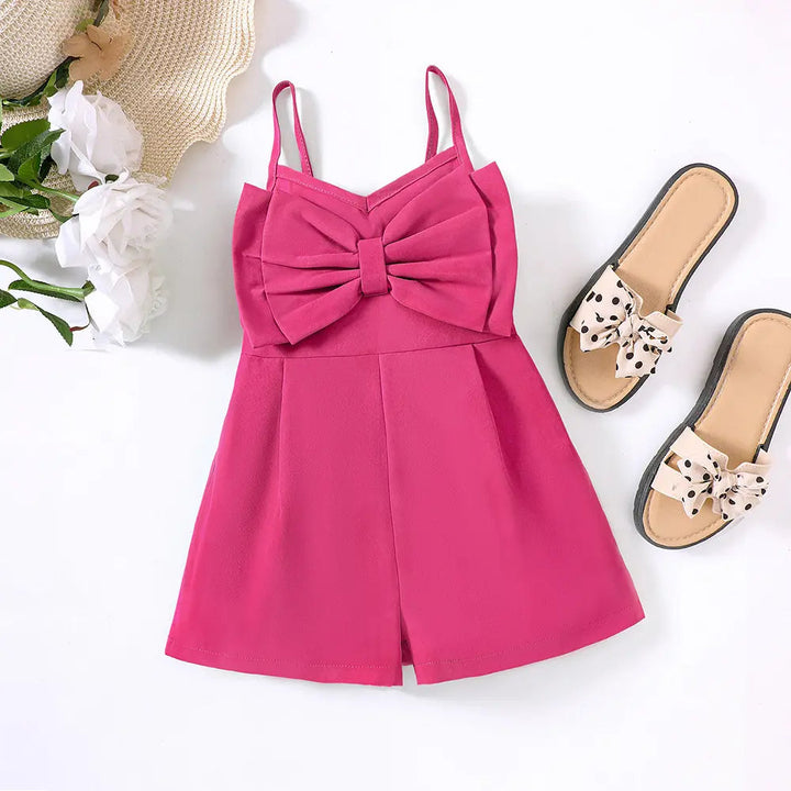 Bright pink bow-front romper laid flat beside sandals, white flowers and a straw hat