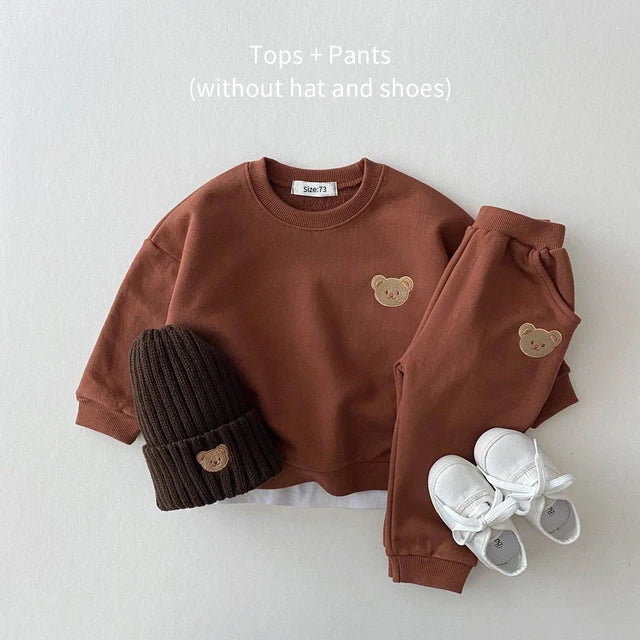 Brown sweatshirt and elastic-waist pants with bear patches, arranged with a dark hat and white shoes