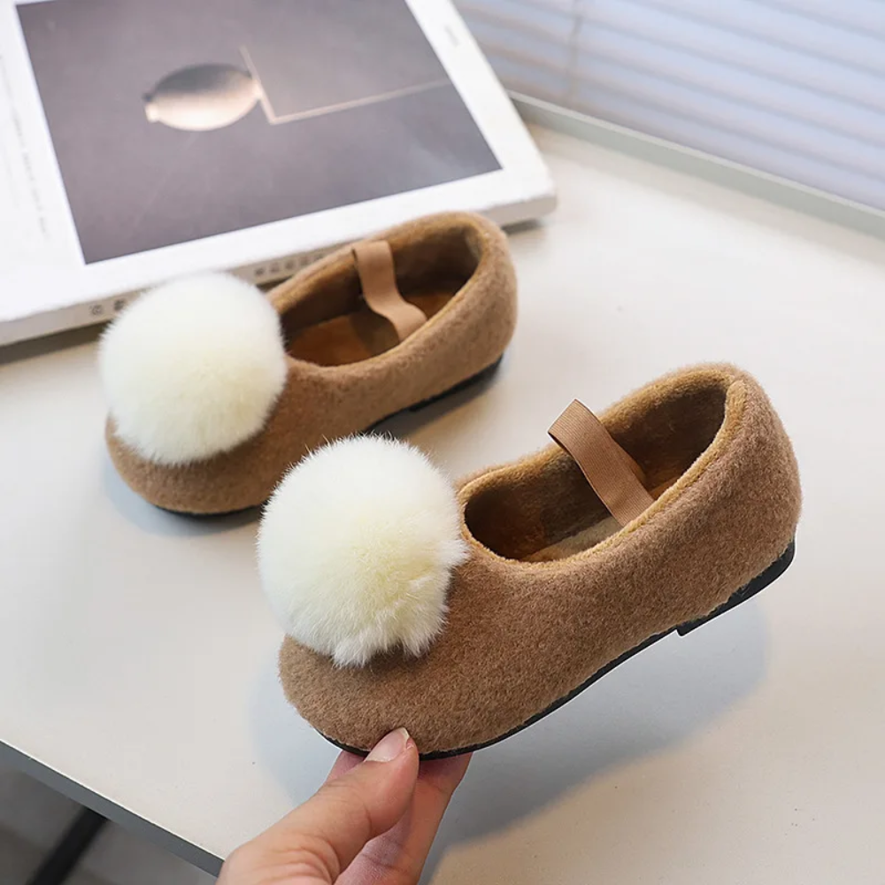 Brown ballet flats with elastic straps and large cream-colored pom-poms