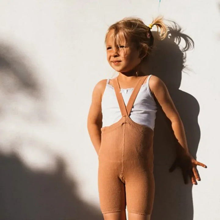 Child wearing brown ribbed suspender shorts with a white top beside a wall.