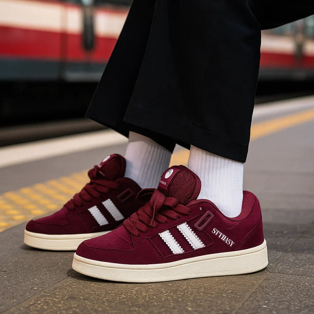 Burgundy low-top sneakers worn with white socks and black pants on a station platform