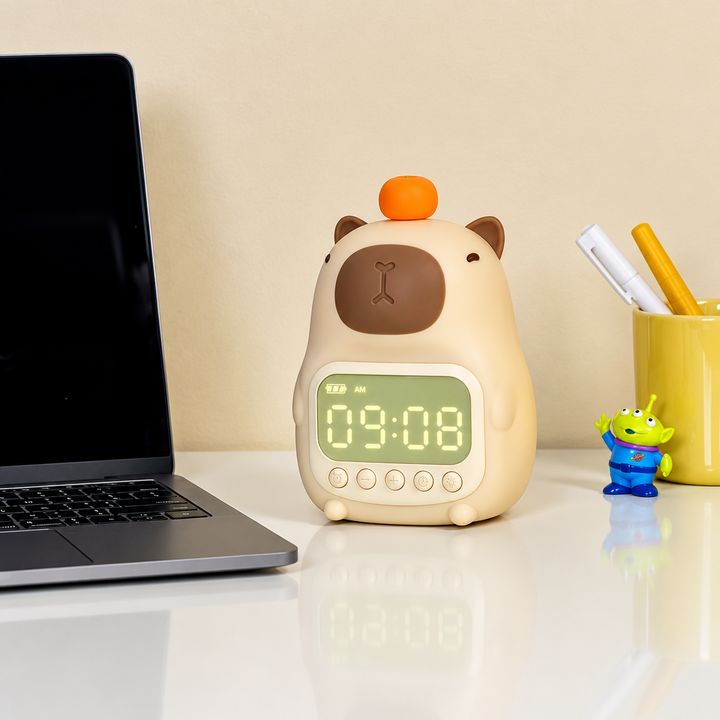 Cream-colored capybara digital clock beside a laptop and pencil cup on a desk.