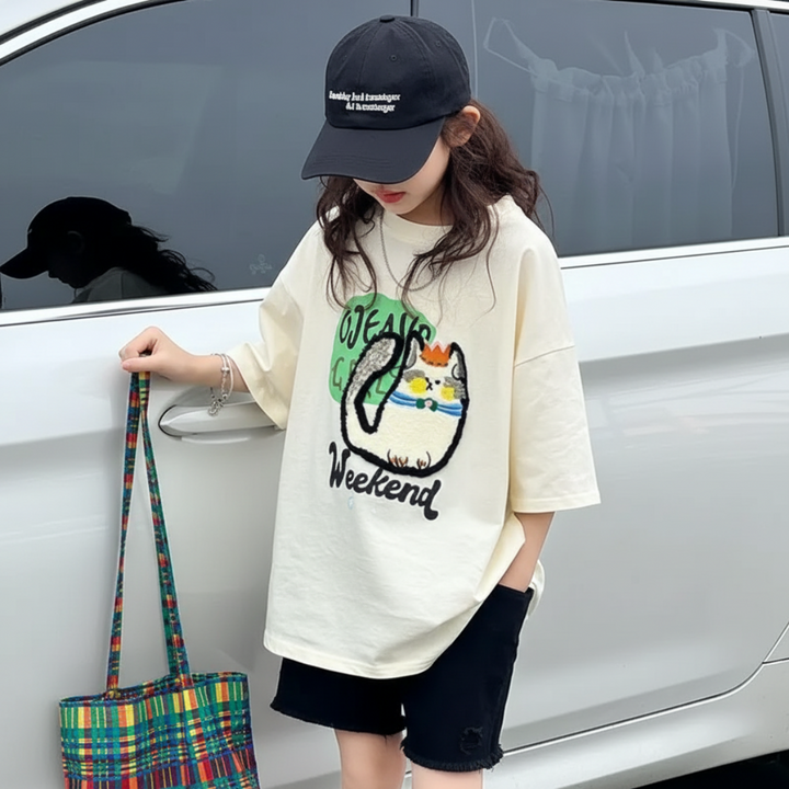 Child wearing the cream cat-print T-shirt beside a car while holding a plaid bag