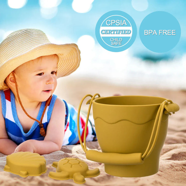Child sitting on the sand beside a green silicone bucket and yellow sea-themed molds.