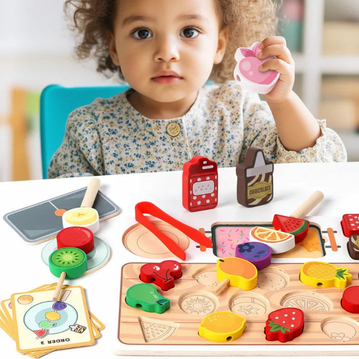 Child holding a pink fruit piece beside the wooden sorting board, skewers and order cards