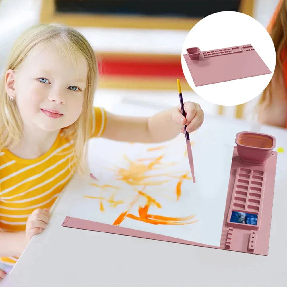 Child painting on paper over a pink craft mat with paint wells and a water cup.